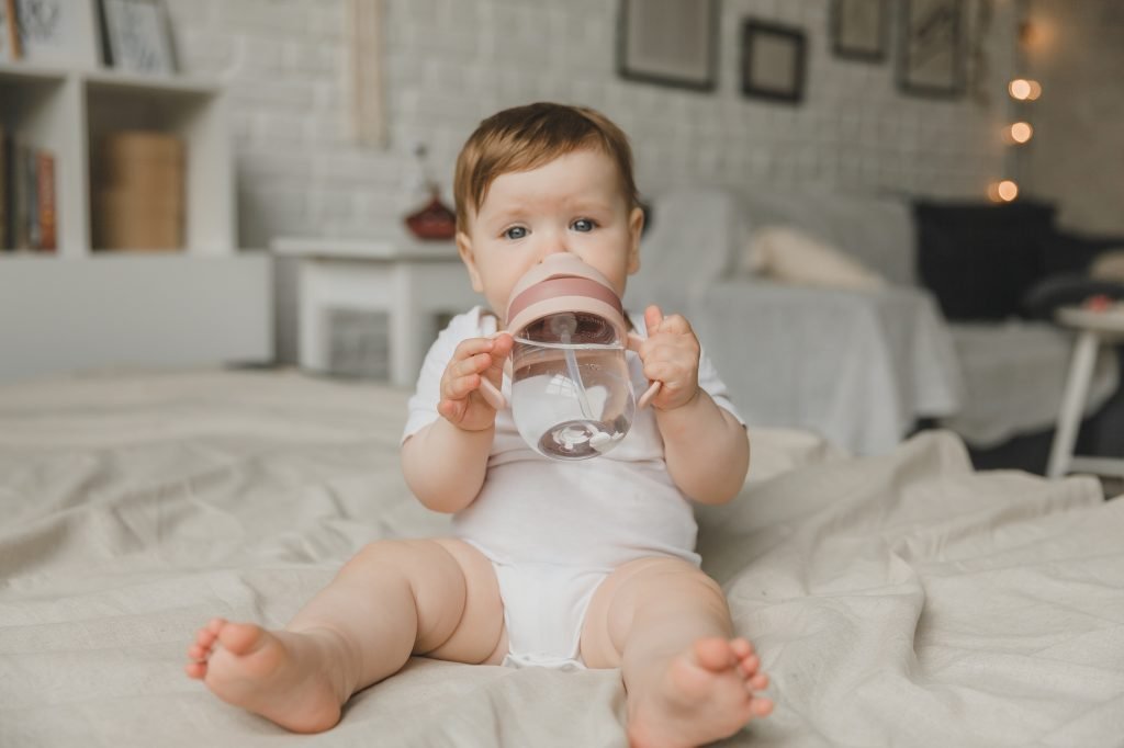 A baby with big blue eyes on a white bed with a special bottle of water with a pacifier.