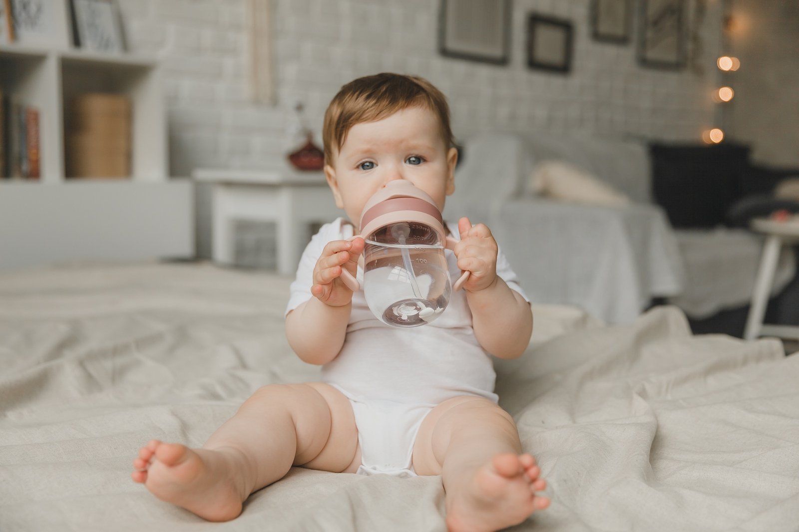 A baby with big blue eyes on a white bed with a special bottle of water with a pacifier.